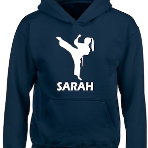 Puede incluir: Una sudadera con capucha azul marino que presenta una silueta gr&aacute;fica blanca de una persona realizando una patada de artes marciales. Debajo del gr&aacute;fico, el nombre SARAH est&aacute; impreso en letras may&uacute;sculas blancas. Incluye un bolsillo delantero.