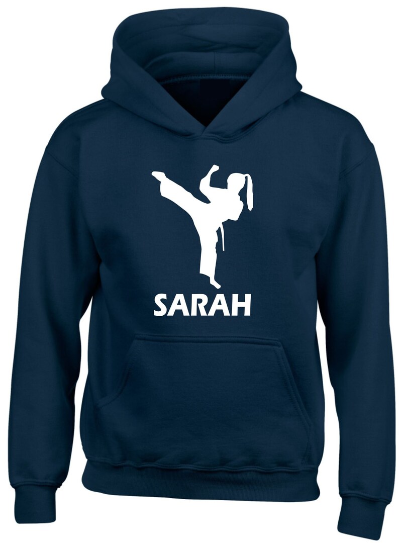 Puede incluir: Una sudadera con capucha azul marino que presenta una silueta gr&aacute;fica blanca de una persona realizando una patada de artes marciales. Debajo del gr&aacute;fico, el nombre SARAH est&aacute; impreso en letras may&uacute;sculas blancas. Incluye un bolsillo delantero.