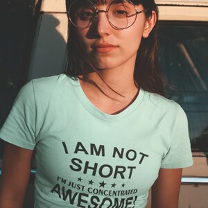 Puede incluir: Camiseta azul claro con el texto negro "I AM NOT SHORT I'M JUST CONCENTRATED AWESOME!". La persona de la imagen lleva gafas redondas y una falda estampada.