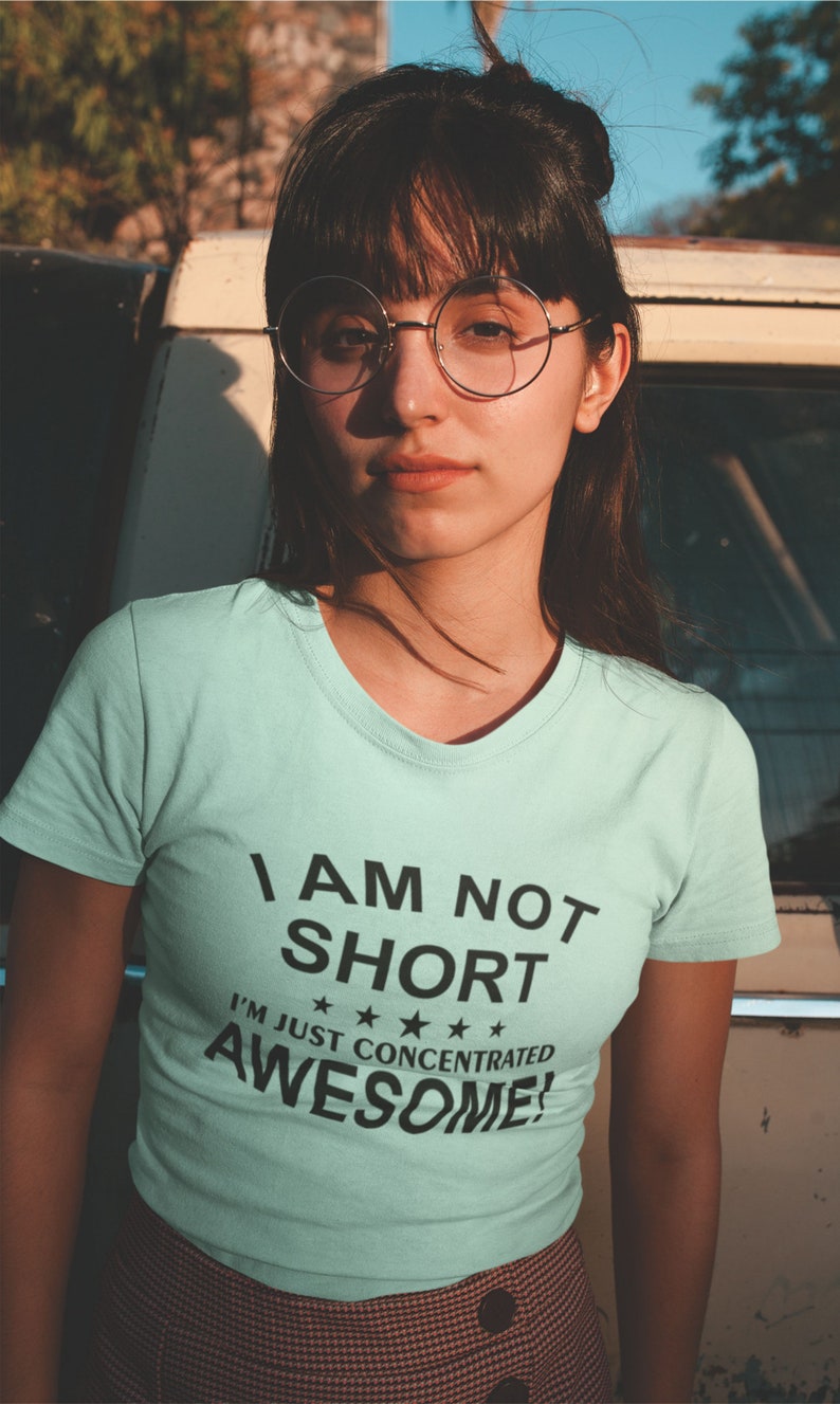 Puede incluir: Camiseta azul claro con el texto negro "I AM NOT SHORT I'M JUST CONCENTRATED AWESOME!". La persona de la imagen lleva gafas redondas y una falda estampada.
