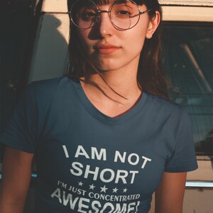 Puede incluir: Una mujer con una camiseta azul con cuello en V con el texto "I AM NOT SHORT I'M JUST CONCENTRATED AWESOME!" en blanco. Lleva gafas redondas y tiene el pelo oscuro. El fondo es un veh&iacute;culo vintage.
