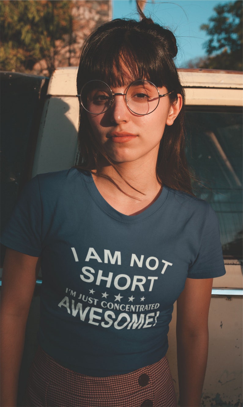 Puede incluir: Una mujer con una camiseta azul con cuello en V con el texto "I AM NOT SHORT I'M JUST CONCENTRATED AWESOME!" en blanco. Lleva gafas redondas y tiene el pelo oscuro. El fondo es un veh&iacute;culo vintage.