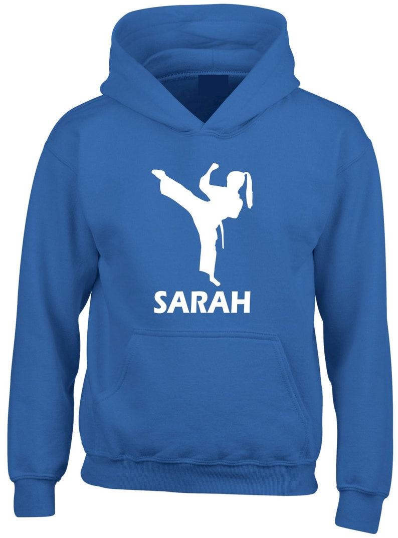 Puede incluir: Una sudadera con capucha de color azul brillante que presenta una silueta gr&aacute;fica blanca de una persona realizando una patada de artes marciales. Debajo del gr&aacute;fico, el nombre SARAH est&aacute; impreso en letras may&uacute;sculas blancas. Bolsillo frontal.