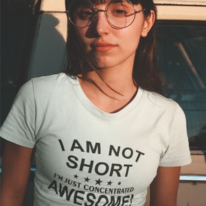 Puede incluir: Camiseta blanca con el texto "I AM NOT SHORT I'M JUST CONCENTRATED AWESOME!" en negro. La persona lleva gafas redondas con montura dorada y una falda estampada. La imagen est&aacute; tomada al aire libre.
