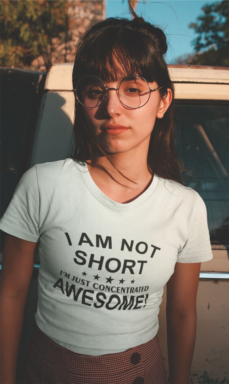 Puede incluir: Camiseta blanca con el texto "I AM NOT SHORT I'M JUST CONCENTRATED AWESOME!" en negro. La persona lleva gafas redondas con montura dorada y una falda estampada. La imagen est&aacute; tomada al aire libre.