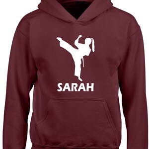 Puede incluir: Una sudadera con capucha de color granate que presenta una silueta gr&aacute;fica blanca de una persona realizando una patada alta con uniforme de artes marciales. Debajo del gr&aacute;fico, el nombre SARAH est&aacute; impreso en letras may&uacute;sculas blancas.