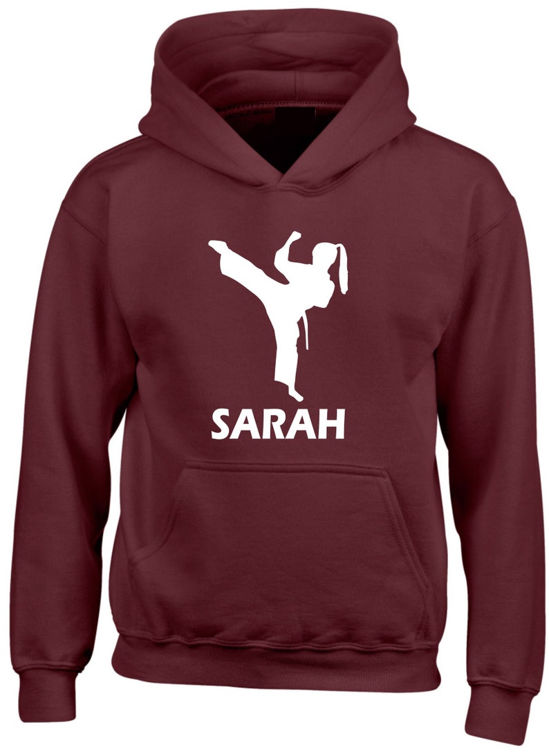 Puede incluir: Una sudadera con capucha de color granate que presenta una silueta gr&aacute;fica blanca de una persona realizando una patada alta con uniforme de artes marciales. Debajo del gr&aacute;fico, el nombre SARAH est&aacute; impreso en letras may&uacute;sculas blancas.
