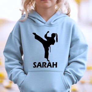 Puede incluir: Una sudadera con capucha de color azul claro que presenta una silueta gr&aacute;fica negra de una persona realizando una patada de artes marciales. Debajo de la silueta, el nombre SARAH est&aacute; impreso en letras may&uacute;sculas negras. Bolsillo frontal.