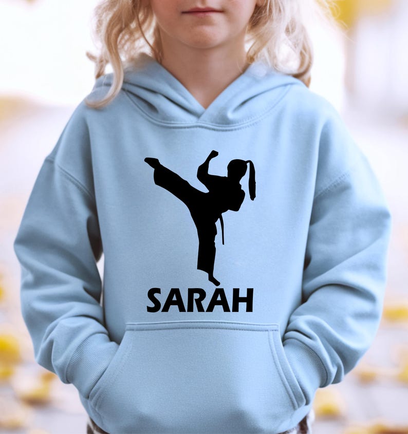 Puede incluir: Una sudadera con capucha de color azul claro que presenta una silueta gr&aacute;fica negra de una persona realizando una patada de artes marciales. Debajo de la silueta, el nombre SARAH est&aacute; impreso en letras may&uacute;sculas negras. Bolsillo frontal.