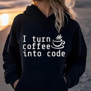 May include: A black long-sleeve hooded sweatshirt featuring white text that reads I turn coffee into code, accompanied by a graphic of a steaming coffee cup. The garment includes a front kangaroo pocket and a hood with matching drawstrings.