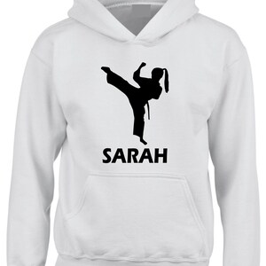 Puede incluir: Una sudadera con capucha blanca que presenta un gr&aacute;fico de silueta negra de una persona realizando una patada alta con uniforme de artes marciales. Debajo del gr&aacute;fico, el nombre SARAH est&aacute; impreso en letras may&uacute;sculas negras y negritas.