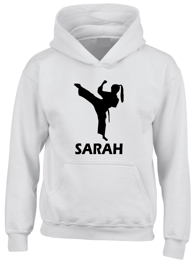 Puede incluir: Una sudadera con capucha blanca que presenta un gr&aacute;fico de silueta negra de una persona realizando una patada alta con uniforme de artes marciales. Debajo del gr&aacute;fico, el nombre SARAH est&aacute; impreso en letras may&uacute;sculas negras y negritas.