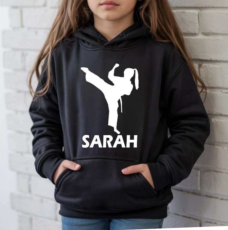 Puede incluir: Una sudadera con capucha negra que presenta una silueta gr&aacute;fica blanca de una persona realizando una patada alta con uniforme de artes marciales. Debajo del gr&aacute;fico, el nombre SARAH est&aacute; impreso en letras may&uacute;sculas blancas. Bolsillo frontal.