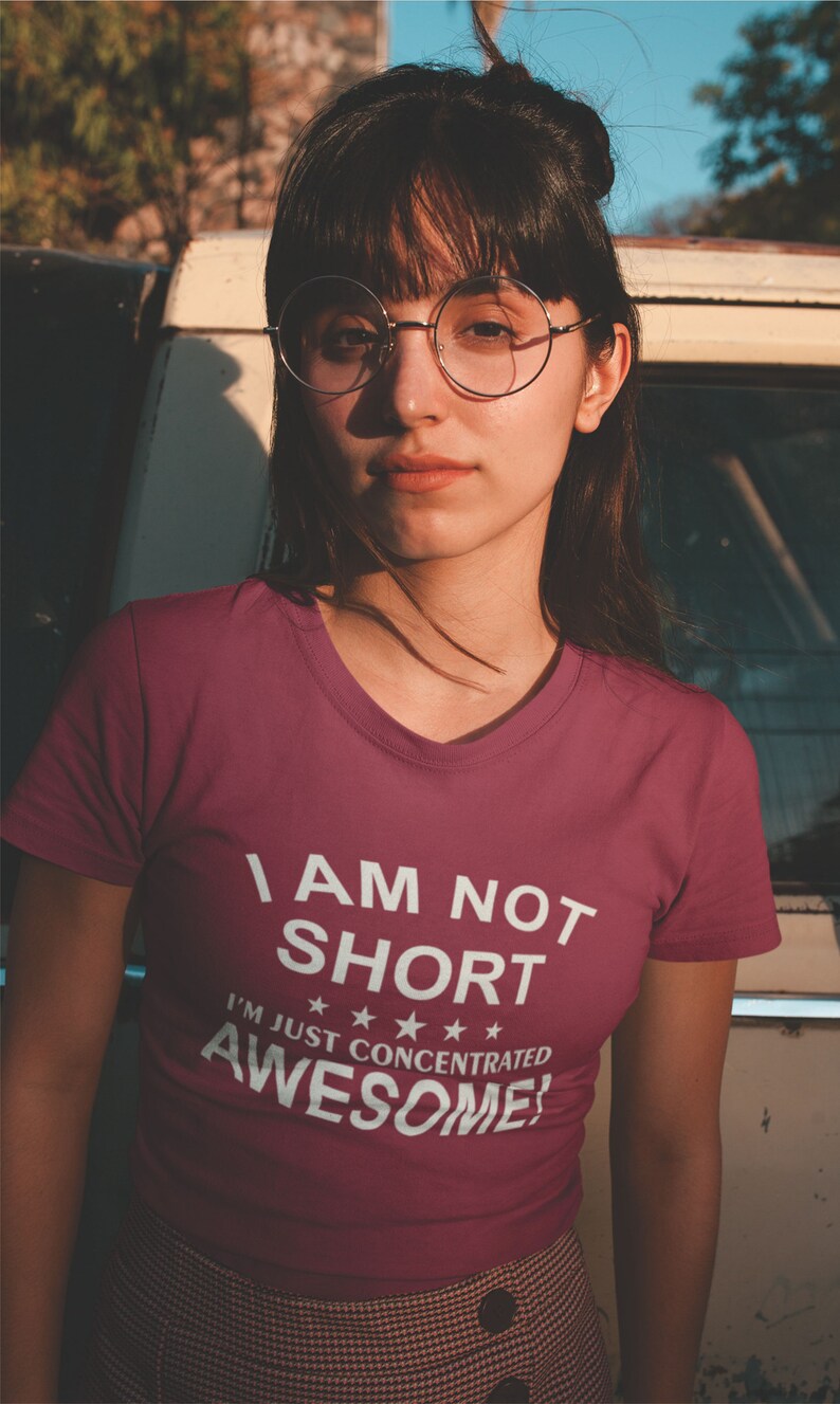 Puede incluir: Camiseta burdeos con cuello en V con el texto blanco "I AM NOT SHORT I'M JUST CONCENTRATED AWESOME!". La persona lleva gafas redondas.
