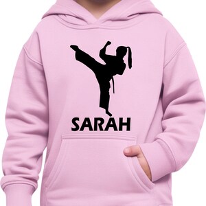 Puede incluir: Una sudadera con capucha de color rosa claro que presenta un gr&aacute;fico de silueta negra de una persona realizando una patada de artes marciales. Debajo del gr&aacute;fico, el nombre SARAH est&aacute; impreso en letras may&uacute;sculas negras. Incluye un bolsillo frontal.