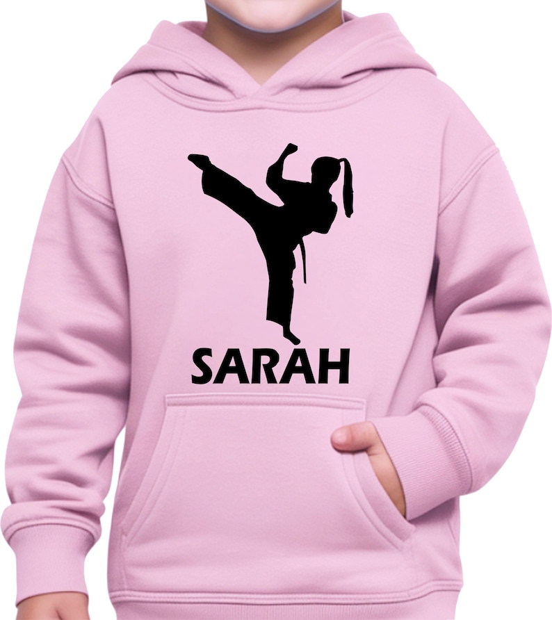 Puede incluir: Una sudadera con capucha de color rosa claro que presenta un gr&aacute;fico de silueta negra de una persona realizando una patada de artes marciales. Debajo del gr&aacute;fico, el nombre SARAH est&aacute; impreso en letras may&uacute;sculas negras. Incluye un bolsillo frontal.