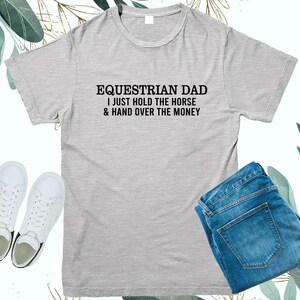 May include: Gray t-shirt with the text "Equestrian Dad I just hold the horse & hand over the money" printed in black.