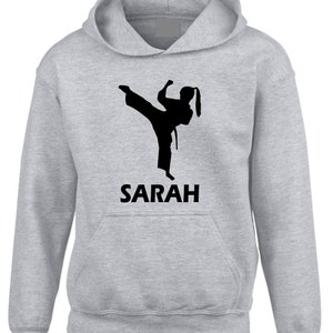 Puede incluir: Una sudadera con capucha de color gris claro que presenta un gr&aacute;fico de silueta negra de una persona realizando una patada alta con uniforme de artes marciales. Debajo del gr&aacute;fico, el nombre SARAH est&aacute; impreso en letras may&uacute;sculas negras.