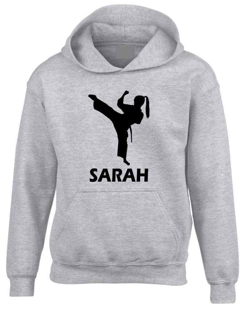 Puede incluir: Una sudadera con capucha de color gris claro que presenta un gr&aacute;fico de silueta negra de una persona realizando una patada alta con uniforme de artes marciales. Debajo del gr&aacute;fico, el nombre SARAH est&aacute; impreso en letras may&uacute;sculas negras.