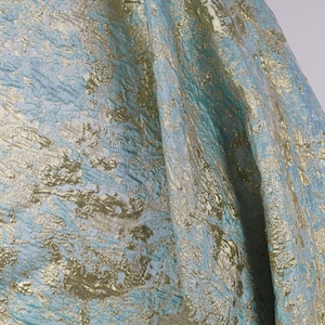 Könnte beinhalten: Nahaufnahme eines strukturierten Stoffes mit schimmerndem Effekt. Der Stoff weist eine Mischung aus hellblauen, teal- und goldfarbenen Farben auf, die ein luxuriöses und elegantes Aussehen erzeugt. Das Material hat einen fließenden Fall.