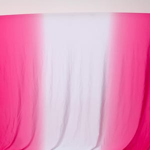 Könnte beinhalten: Ein Stoff in Pink und Weiß mit Farbverlauf, drapiert über einem Holzrahmen. Der Stoff wechselt von hellem Pink an den Rändern zu Weiß in der Mitte. Das Material wirkt weich und fließend, geeignet für verschiedene Zwecke.