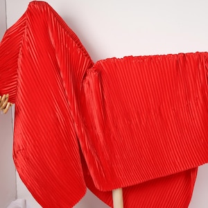 May include: Vibrant red pleated fabric, displayed on a wooden rod and a mannequin arm. The fabric is arranged to resemble a garment, with vertical pleats creating a textured surface. The overall effect is a display of bright, flowing material.