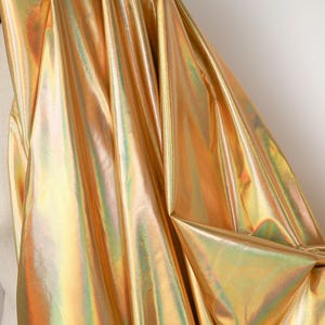 Può includere: Un tessuto drappeggiato con una finitura dorata scintillante e iridescente. Il materiale ha una superficie liscia e riflettente con sottili variazioni di colore, creando un effetto visivo dinamico. Il tessuto è probabilmente utilizzato per abbigliamento o scopi decorativi.