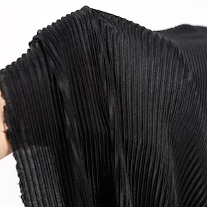 May include: Close-up of black pleated fabric. The material has vertical pleats and a slightly shiny appearance. The fabric is held up, showing the texture and drape. The background is plain white.