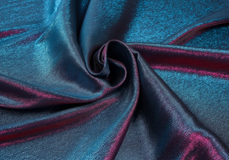 May include: Close-up of iridescent fabric with a blue and purple color gradient. The fabric is folded and draped, showing its texture and sheen.