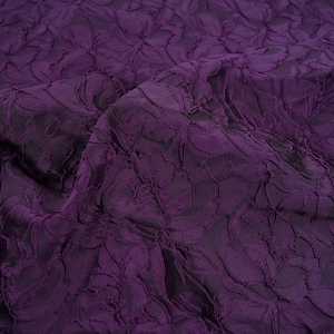 Pu&ograve; includere: Primo piano di un tessuto viola scuro con un motivo floreale in rilievo. Il tessuto ha un aspetto strutturato e tridimensionale, con il design floreale che crea ombre e riflessi. Il materiale &egrave; drappeggiato, mostrando pieghe e movimento.