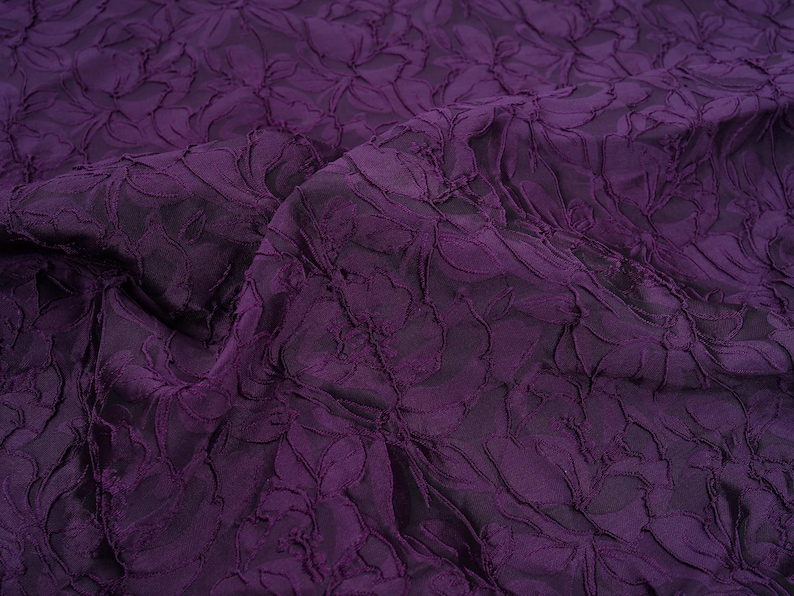 Pu&ograve; includere: Primo piano di un tessuto viola scuro con un motivo floreale in rilievo. Il tessuto ha un aspetto strutturato e tridimensionale, con il design floreale che crea ombre e riflessi. Il materiale &egrave; drappeggiato, mostrando pieghe e movimento.