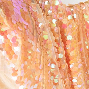 May include: Close-up of a fabric covered in iridescent sequins. The sequins are primarily peach and orange, with hints of pink and white. The fabric is draped, creating folds and shadows, and is likely used for apparel or decorative purposes.
