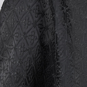 Può includere: Tessuto nero con un motivo floreale in rilievo ripetuto. Il design presenta forme a diamante con fiori a quattro petali al centro e agli angoli. Il tessuto è drappeggiato, mostrando la consistenza e la profondità del design.