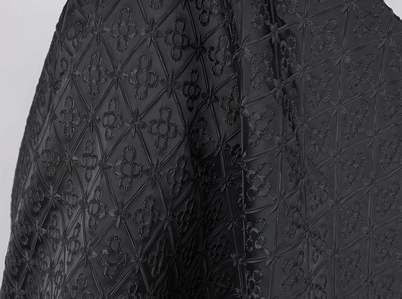 K&ouml;nnte beinhalten: Schwarzer Stoff mit einem sich wiederholenden gepr&auml;gten Blumenmuster. Das Design zeigt Rauten mit vierbl&auml;ttrigen Blumen in der Mitte und in den Ecken. Der Stoff ist drapiert und zeigt die Textur und Tiefe des Designs.