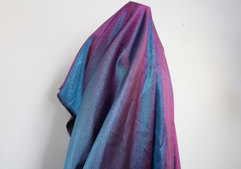 May include: A shimmering iridescent fabric with a blue and purple gradient. The fabric is draped over a white surface.
