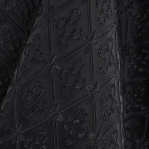 K&ouml;nnte beinhalten: Nahaufnahme eines schwarzen Stoffes mit einem sich wiederholenden Muster aus stilisierten floralen und geometrischen Formen. Der Stoff hat eine strukturierte Oberfl&auml;che, wobei das Design leicht erhaben ist. Der Gesamteffekt ist elegant und anspruchsvoll.