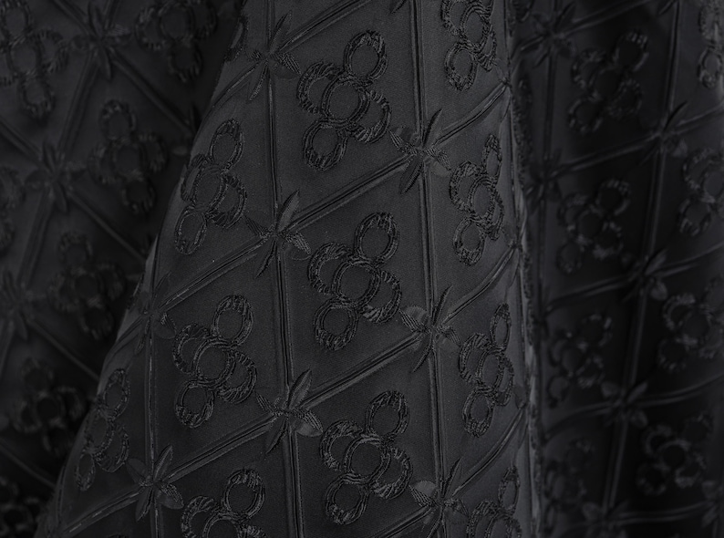 K&ouml;nnte beinhalten: Nahaufnahme eines schwarzen Stoffes mit einem sich wiederholenden Muster aus stilisierten floralen und geometrischen Formen. Der Stoff hat eine strukturierte Oberfl&auml;che, wobei das Design leicht erhaben ist. Der Gesamteffekt ist elegant und anspruchsvoll.