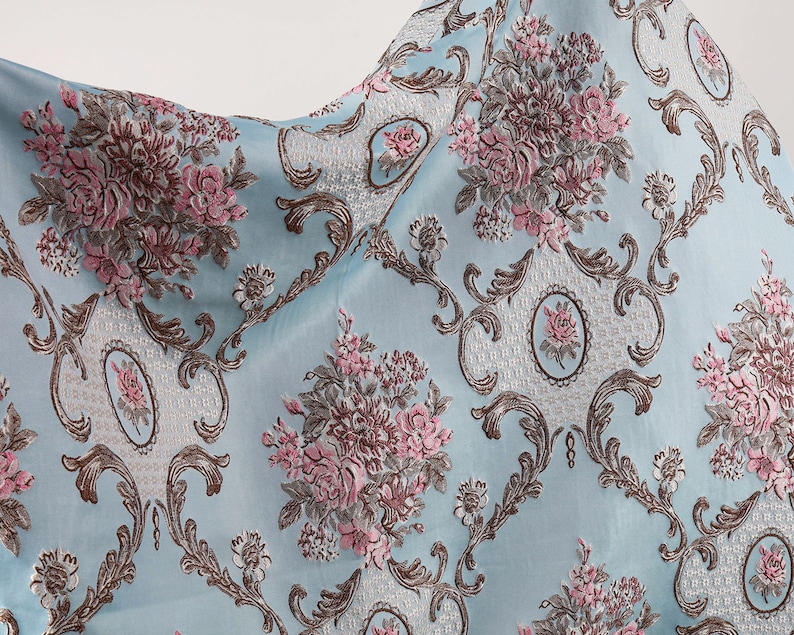 May include: A light blue fabric with a floral pattern. The fabric features a repeating design of pink and brown flowers with intricate gold scrollwork.