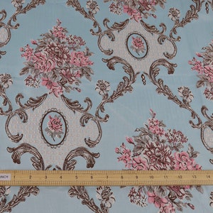 May include: A close-up of a light blue fabric with a repeating pattern of pink and brown floral bouquets and ornate scrollwork. A ruler is placed at the bottom of the image, showing the fabric is 16 inches wide.
