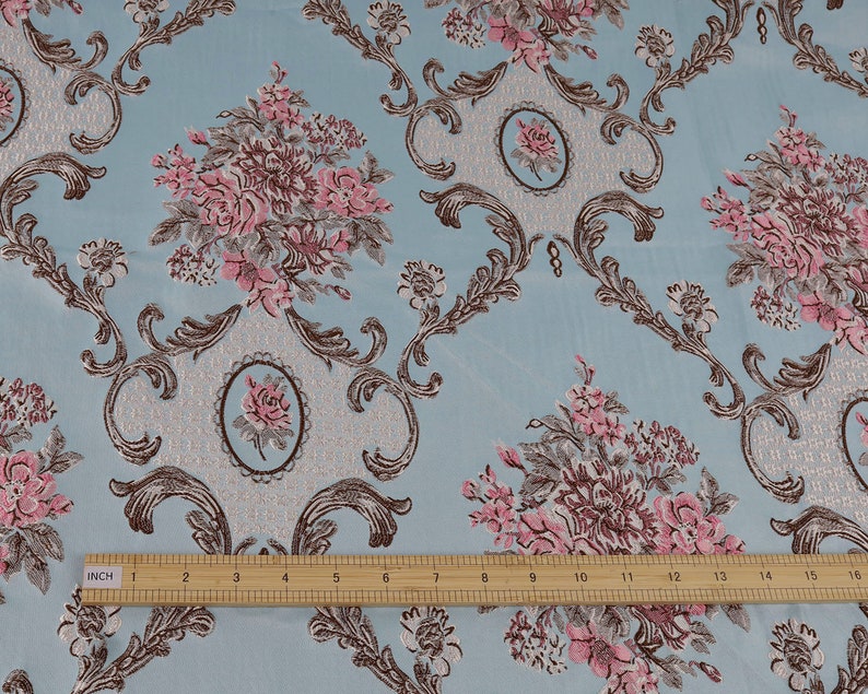 May include: A close-up of a light blue fabric with a repeating pattern of pink and brown floral bouquets and ornate scrollwork. A ruler is placed at the bottom of the image, showing the fabric is 16 inches wide.