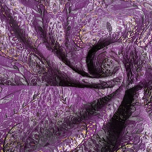 May include: Close-up of a luxurious purple fabric with a detailed paisley pattern. The fabric has a textured appearance with silver and gold accents, creating a rich and elegant look. The fabric is draped, showing its folds and texture.