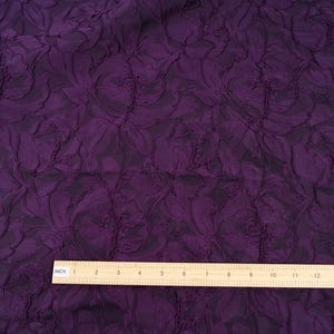 Pu&ograve; includere: Primo piano di un tessuto viola scuro con un motivo floreale in rilievo. Il tessuto &egrave; strutturato e ha uno sfondo scuro. Un righello di legno, segnato in pollici, &egrave; posto sul fondo del tessuto.