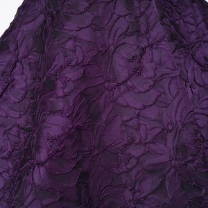 Pu&ograve; includere: Primo piano di un tessuto viola scuro con un motivo floreale. Il tessuto presenta un design strutturato e in rilievo di grandi fiori e foglie. Il colore &egrave; una ricca e scura tonalit&agrave; di viola, con sfumature di nero sullo sfondo.