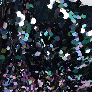 May include: Close-up of a fabric covered in iridescent sequins. The sequins are primarily black, with hints of green, blue, and purple. The fabric appears to be draped, with a black mesh backing visible.