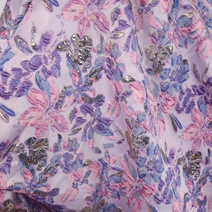 Puede incluir: Primer plano de una tela con un estampado floral. El diseño presenta grandes flores en tonos lavanda, rosa y morado, con detalles plateados metálicos. La tela tiene una apariencia suave y fluida, lo que sugiere que podría usarse para prendas de vestir o artículos decorativos.
