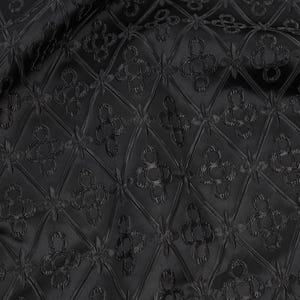K&ouml;nnte beinhalten: Schwarzer Stoff mit einem sich wiederholenden Muster aus Rauten und floralen Motiven. Der Stoff hat einen leichten Glanz, und das Muster ist erhaben, wodurch eine strukturierte Oberfl&auml;che entsteht. Der Gesamteffekt ist elegant und raffiniert.