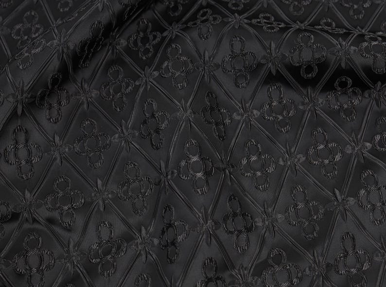 K&ouml;nnte beinhalten: Schwarzer Stoff mit einem sich wiederholenden Muster aus Rauten und floralen Motiven. Der Stoff hat einen leichten Glanz, und das Muster ist erhaben, wodurch eine strukturierte Oberfl&auml;che entsteht. Der Gesamteffekt ist elegant und raffiniert.