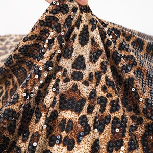Puede incluir: Primer plano de una tela con estampado de leopardo. La tela presenta un patrón de manchas marrones y negras sobre un fondo beige. Pequeñas lentejuelas brillantes están dispersas por la superficie, añadiendo un toque de brillo. La tela se sostiene, mostrando su caída.
