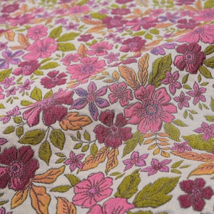 Puede incluir: Primer plano de una tela floral con un patrón repetido de flores rosas, magenta y moradas, junto con hojas verdes y naranjas. La tela tiene una apariencia texturizada y un fondo neutro.