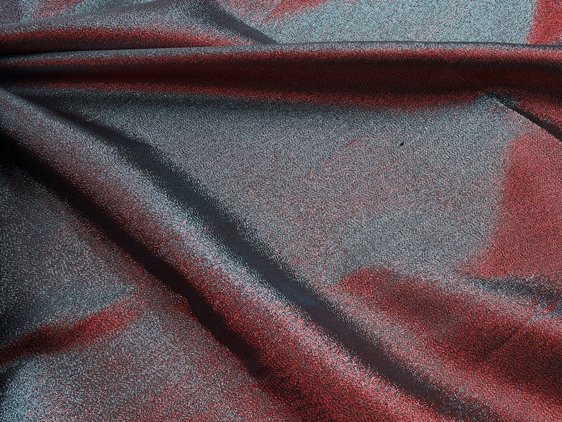 May include: Close-up of a shimmering fabric with a red and silver color gradient. The fabric is folded and draped, creating soft folds and wrinkles.